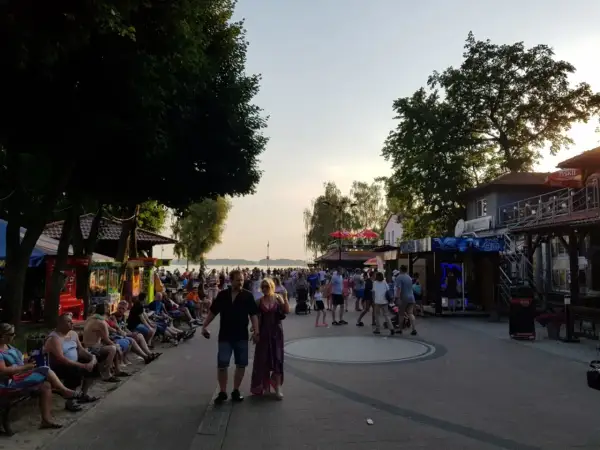 Tłum ludzi spaceruje promenadą w Skorzęcinie, korzystając z licznych atrakcji i letniej atmosfery nad jeziorem.