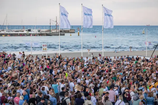 Tłumy ludzi na plaży w Sopocie, cieszące się letnimi imprezami. W tle morze i port.