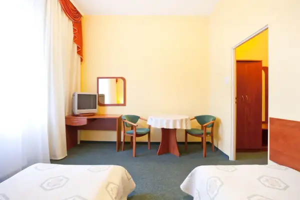 Pokój hotelowy z dwoma łóżkami, biurkiem z telewizorem, stolikiem z krzesłami i szafą.