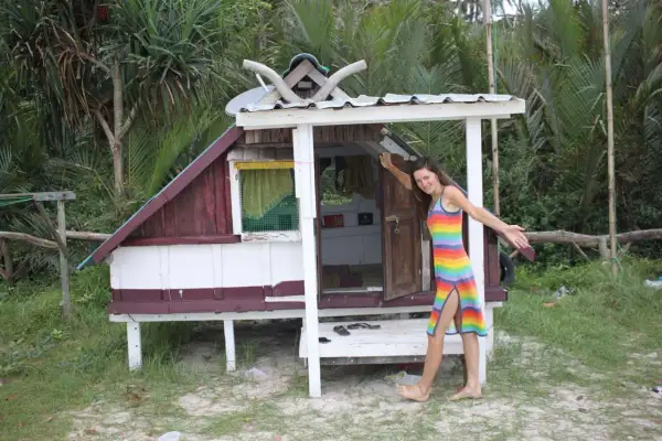 Kobieta w tęczowej sukience stoi obok małego, drewnianego domku na plaży.