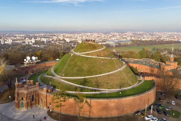 Kopiec Kościuszki, zielona kopuła z krętymi ścieżkami, góruje nad ceglanymi fortami i rozległym, **starym miastem w Krakowie**.